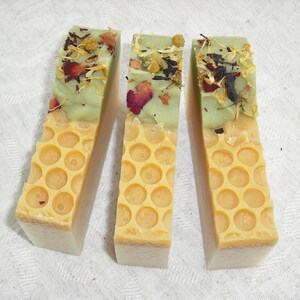 May include: Three bars of handmade soap with a honeycomb design. The top layer of each bar is green and has dried flowers and herbs embedded in it. The bottom layer is a light yellow color.