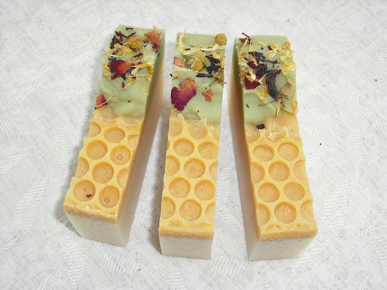 May include: Three bars of handmade soap with a honeycomb design. The top layer of each bar is green and has dried flowers and herbs embedded in it. The bottom layer is a light yellow color.