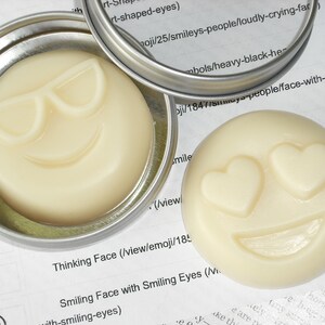 May include: Two small round containers with lids, each containing a white, solid, round soap with a different smiley face design. The first container has a soap with sunglasses and a smile. The second container has a soap with heart-shaped eyes and a smile. The text on the label reads "Thinking Face (view/emoji/18..." and "Smiling Face with Smiling Eyes (Vi..."