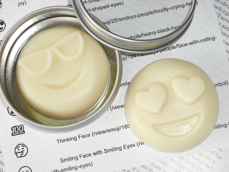May include: Two small round containers with lids, each containing a white, solid, round soap with a different smiley face design. The first container has a soap with sunglasses and a smile. The second container has a soap with heart-shaped eyes and a smile. The text on the label reads "Thinking Face (view/emoji/18..." and "Smiling Face with Smiling Eyes (Vi..."