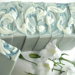 May include: A series of rectangular soap bars featuring a solid grey base and a decorative top layer with a marbled swirl pattern in shades of white and light blue. The soap bars are arranged in a row, accompanied by white bell-shaped flowers.