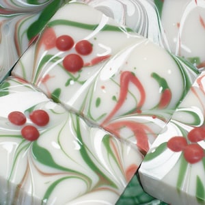 May include: Close-up of several square bars of soap with a festive design. The soap features a white base with swirls of green and red, and small red spheres. The design evokes a holiday theme.