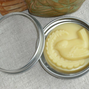 May include: A small round tin container with a silver lid. The lid is open and the container holds a pale yellow, bird-shaped, scented candle.