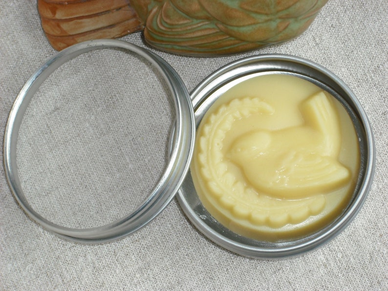May include: A small round tin container with a silver lid. The lid is open and the container holds a pale yellow, bird-shaped, scented candle.