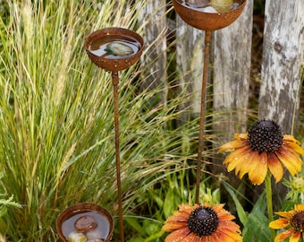 May include: Three rustic metal bird baths with water and stones, set in a garden with tall grass and orange coneflowers. The bird baths have a weathered, rusty appearance and are on slender stems.