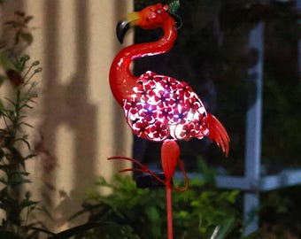 Könnte beinhalten: Ein roter Metall-Flamingoständer für den Garten mit solarbetriebenen Lichtern. Der Flamingo ist mit roten Blumen verziert und hat ein weißes Licht, das durch die Blumen scheint.