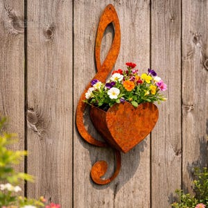 Puede incluir: Una jardinera de metal oxidado en forma de clave de sol con una maceta en forma de corazón. La jardinera está llena de flores coloridas contra una valla de madera desgastada. Las flores incluyen flores blancas, amarillas, moradas y rojas.