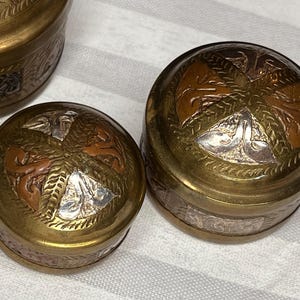 May include: Three small, round, decorative brass boxes with lids. The boxes have intricate patterns in silver and copper accents. The lids feature a central design with floral and geometric elements. The boxes are likely for storing small items.
