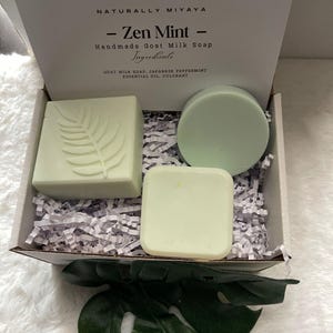 May include: A gift box filled with three bars of green goat milk soap. The soap bars are in a variety of shapes, including a square, a rectangle, and a circle. The box is lined with white shredded paper. The soap is labeled "Zen Mint" and lists the ingredients as "Goat Milk Soap, Japanese Peppermint Essential Oil, Colorant".