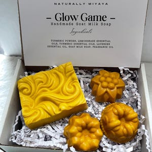 May include: A gift box set of four yellow goat milk soap bars. The soap bars are in the shape of a rectangle, a flower, and two circles. The box is white with the words "Glow Game" and "Handmade Goat Milk Soap" printed on the lid. The ingredients are listed on the lid: "Turmeric Powder, Lemongrass Essential Oils, Turmeric Essential Oils, Lavender Essential Oil, Goat Milk Soap, Fragrance Oil."