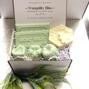 May include: A white gift box filled with green and white goat milk soap bars. The box is decorated with the words "Tranquility Bliss" and "Goat Milk".