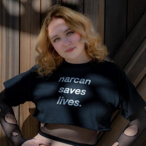 Könnte beinhalten: Ein schwarzes, kurzärmeliges bauchfreies Oberteil mit dem weißen Schriftzug "narcan saves lives." auf der Vorderseite. Das Kleidungsstück wird über einem schwarzen, langärmeligen Netzshirt mit Löchern getragen, kombiniert mit einer blauen Jeans.
