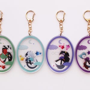 May include: Four oval-shaped keychains with gold-colored clasps. Each keychain features a different character design inside a clear oval frame with a colored border. The designs include stars, moons, and clouds.
