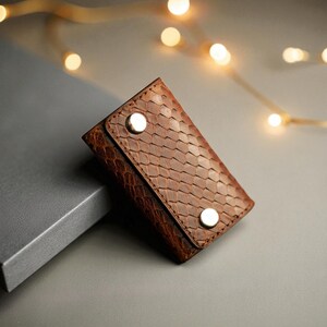 Może przedstawiać: Brązowy skórzany brelok do kluczy z wzorem łusek. Prostokątne etui ma dwa srebrne zatrzaski. Brelok do kluczy jest prezentowany na szarej powierzchni z miękkimi, rozmytymi światłami w tle.