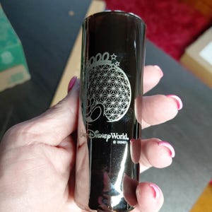 Può includere: Un bicchiere da shot cilindrico nero con un design inciso in argento della struttura Spaceship Earth di Epcot, il testo 2000 e il logo Walt Disney World. La superficie è riflettente e liscia, mostrando un grafico dettagliato della cupola.