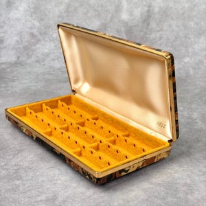 May include: A rectangular jewelry box with a hinged lid, featuring a brown and black mottled exterior pattern. The interior is lined with a smooth cream-colored fabric on the lid and a bright yellow felt insert with slots for rings or earrings.