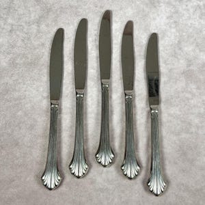 May include: Five silver butter knives with a decorative, ridged handle and a rounded blade. The knives are arranged vertically on a light gray surface. The handles have a fan-like design at the base.