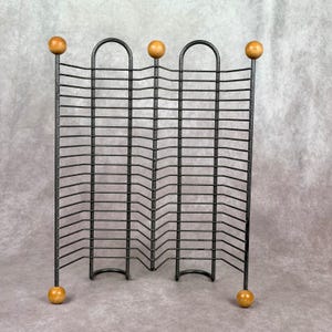 May include: A black metal wire rack featuring a series of horizontal bars arranged in a zig-zag pattern. The structure is supported by five vertical posts, each topped and bottomed with a smooth, round, light-colored wooden ball finial.