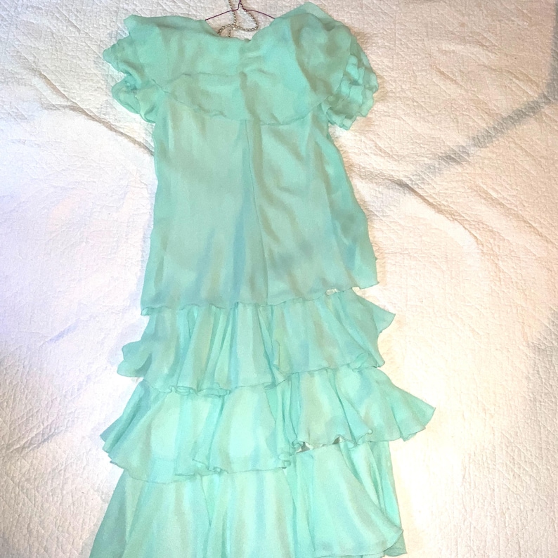 May include: A light turquoise dress with short sleeves and a tiered skirt. The dress is made of a lightweight, flowing fabric and hangs on a hanger. The dress has a ruffled neckline and a layered skirt.