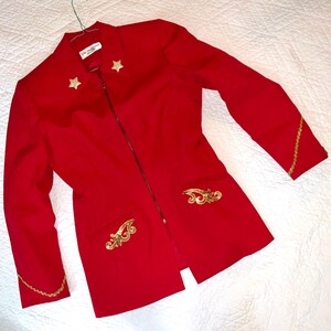 May include: A bright red blazer with a zipper closure, gold star accents, and decorative gold embellishments on the pockets and sleeves. The blazer has a tailored fit and a classic design.