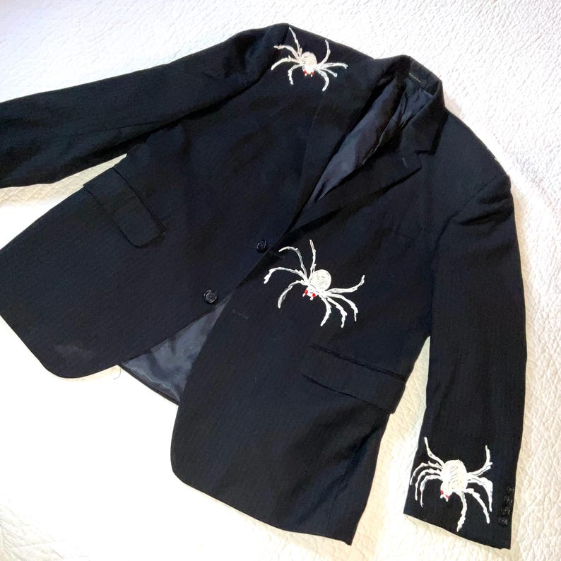 Op de afbeelding: Zwarte blazer met witte spinnenborduurwerk op de borst en mouw. De blazer heeft een krijtstreepmotief en een sluiting met twee knopen. De spinnen hebben rode accenten.