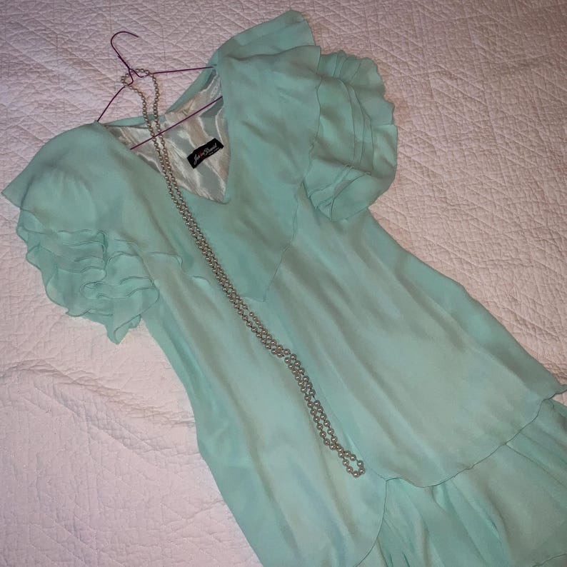 May include: A light turquoise dress with ruffled sleeves and a tiered skirt, hanging on a purple hanger. A long, silver beaded necklace drapes across the dress. The dress has a V-neck and a label.