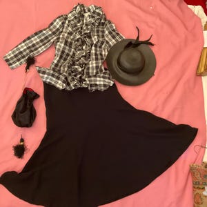 May include: An ensemble featuring a black and white plaid ruffled blouse, a black skirt, a dark hat with feathers, and a small black drawstring bag. The outfit is arranged on a pink surface, suggesting a vintage or retro style.