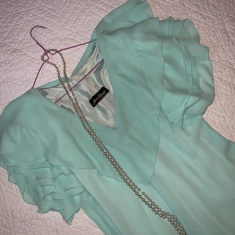 May include: A mint green dress with ruffled sleeves hangs on a purple hanger. A long, beaded necklace drapes across the dress. The dress has a V-neck and a label that reads "Jason David".