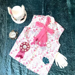 May include: A whimsical costume ensemble featuring a white vest with a floral pattern in pink and blue, a pink scarf, and a decorative rosette. Accessories include a white hat with bunny ears, a pocket watch, a small alarm clock, and white gloves.