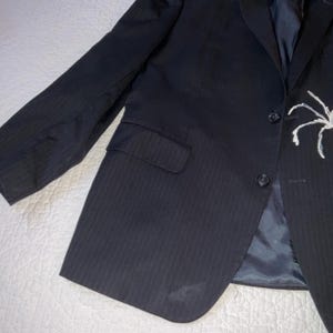 Op de afbeelding: Een donkerblauwe blazer met een subtiel krijtstreepmotief. De blazer heeft een sluiting met twee knopen, een borstzak en een wit spinnendesign op de revers. De jas ligt plat op een witte ondergrond.