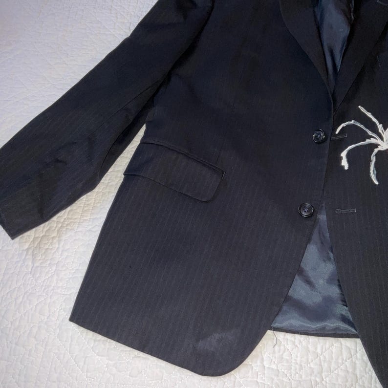 Op de afbeelding: Een donkerblauwe blazer met een subtiel krijtstreepmotief. De blazer heeft een sluiting met twee knopen, een borstzak en een wit spinnendesign op de revers. De jas ligt plat op een witte ondergrond.