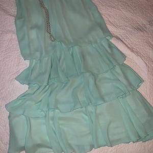 May include: A sleeveless, light turquoise dress with layered ruffles. A delicate pearl necklace drapes across the dress. The dress is made of a flowing, lightweight fabric, perfect for warm weather.