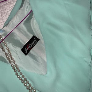 May include: A light turquoise garment with a satin lining and a pearl necklace. The garment has a label that reads "Justin David New York". The fabric has a soft, flowing appearance.