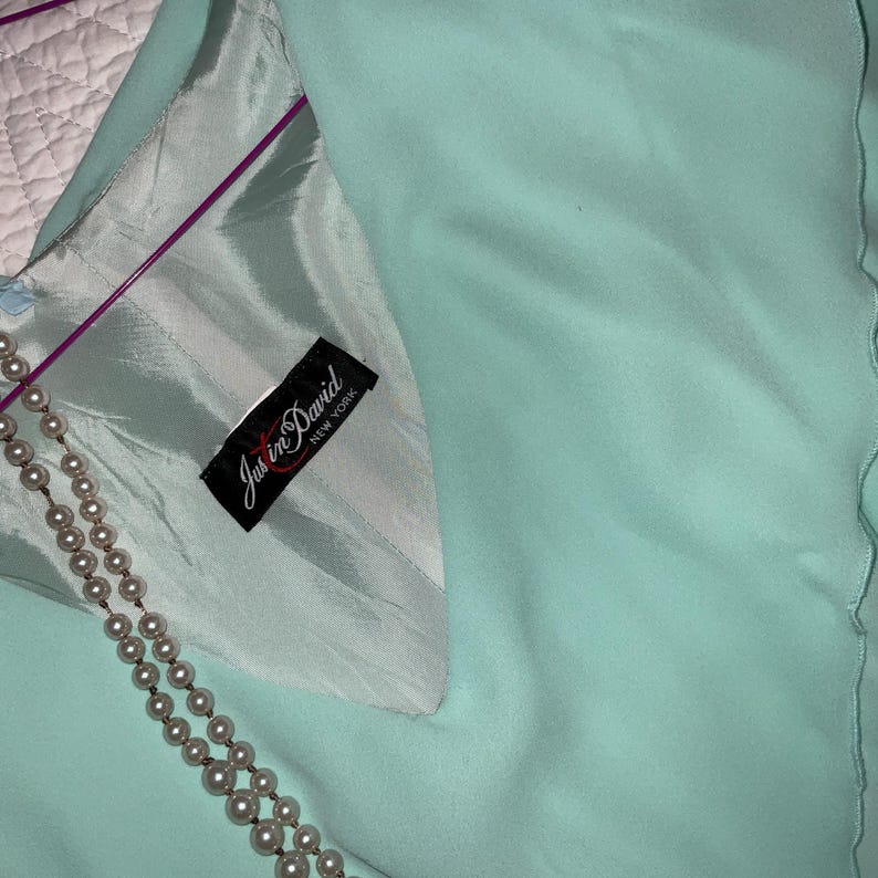 May include: A light turquoise garment with a satin lining and a pearl necklace. The garment has a label that reads "Justin David New York". The fabric has a soft, flowing appearance.