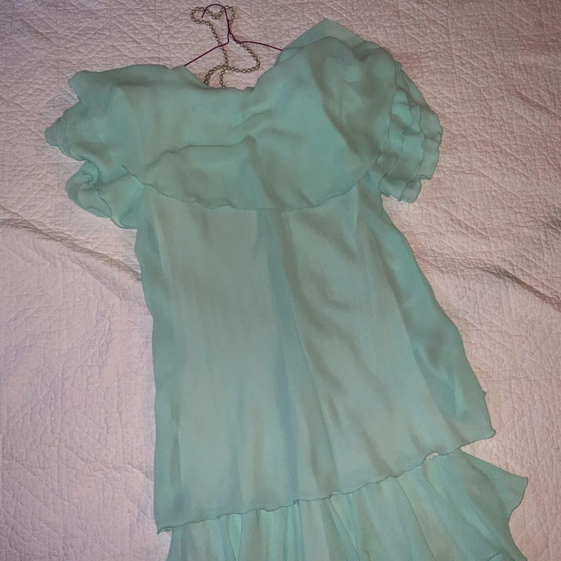 May include: A light turquoise dress with a ruffled neckline and short sleeves hangs on a wire hanger. The dress has a tiered design with a flowing, lightweight fabric. The garment is displayed on a white quilted surface.
