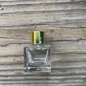 May include: A clear, square glass perfume bottle with a gold-colored cap. The bottle has a rectangular shape with a slightly recessed area. The cap is a shiny gold color. The bottle is sitting on a wooden surface.