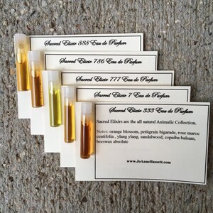 May include: A collection of small glass vials filled with amber and yellow liquids, each labeled with "Sacred Elixir" and a number, such as 888, 786, and 333. The vials are arranged on white cards with product information and the website www.JoAnneBassett.com.