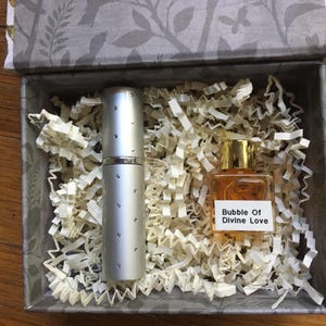 A silver perfume travel atomizer and a 5 ml perfume bottle labeled "Bubble Of Divine Love" sit in a decorative box with shredded paper. The box has a gray floral pattern on the lid and interior.