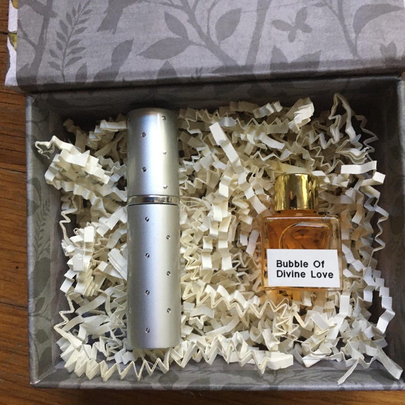 A silver perfume travel atomizer and a 5 ml perfume bottle labeled "Bubble Of Divine Love" sit in a decorative box with shredded paper. The box has a gray floral pattern on the lid and interior.