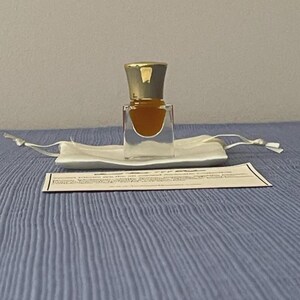 May include: A small, clear glass perfume bottle with a gold-colored cap, filled with a golden-brown liquid. The bottle sits on a white satin pouch, with a small card in front. The background is a light blue surface.