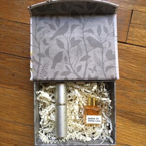 A gift box with a gray bird and floral pattern houses a 5 ml perfume bottle and a silver atomizer. The perfume bottle is labeled "Bubble Of Divine Love." The box is lined with shredded paper.