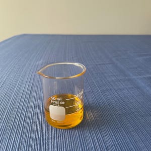 May include: A clear Pyrex beaker containing a yellow liquid. The beaker has a 50 ml capacity and is marked with measurements. The beaker is labeled "PYREX USA" and "No. 1000". The background is a blue textured surface.