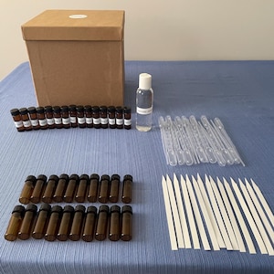 May include: A fragrance kit with a brown cardboard box, small brown glass bottles, a clear bottle, pipettes, and scent strips. The bottles are labeled. The items are arranged on a blue tablecloth.