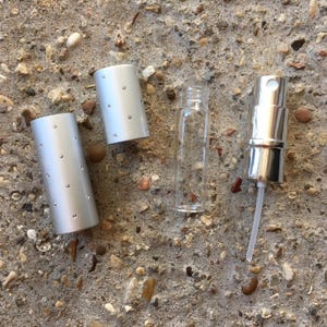 A disassembled perfume travel sprayer. The set includes a clear glass bottle, a silver spray nozzle, and a silver cylindrical cap with small silver dots. The items are arranged on a textured, gray surface.