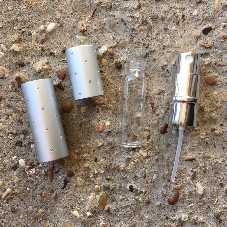A disassembled perfume travel sprayer. The set includes a clear glass bottle, a silver spray nozzle, and a silver cylindrical cap with small silver dots. The items are arranged on a textured, gray surface.