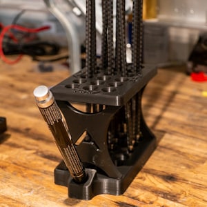 Peut inclure: Un organisateur noir imprimé en 3D avec plusieurs fentes pour ranger des stylos, des crayons ou d'autres ustensiles d'écriture. L'organisateur a un compartiment latéral pour ranger un seul article plus grand, comme un marqueur ou un bâton de colle.