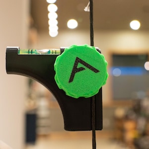 May include: A black level tool with a green circular attachment featuring a black "A" symbol. The tool has a small bubble level and is suspended by a black cord. The background is blurred.