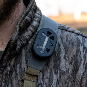 Könnte beinhalten: Eine schwarze und graue Tarnjacke mit einem schwarzen runden Objekt, das an der Schultertrage befestigt ist. Das Objekt hat ein kreisförmiges Design mit einer weißen Mitte.