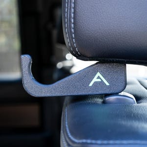 May include: A black plastic hook with a green "A" logo attached to the headrest of a car seat.