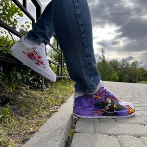 May include: White sneakers with custom artwork. One shoe features pink cherry blossoms, the other a purple and green anime-style design. The shoes are worn with blue jeans.